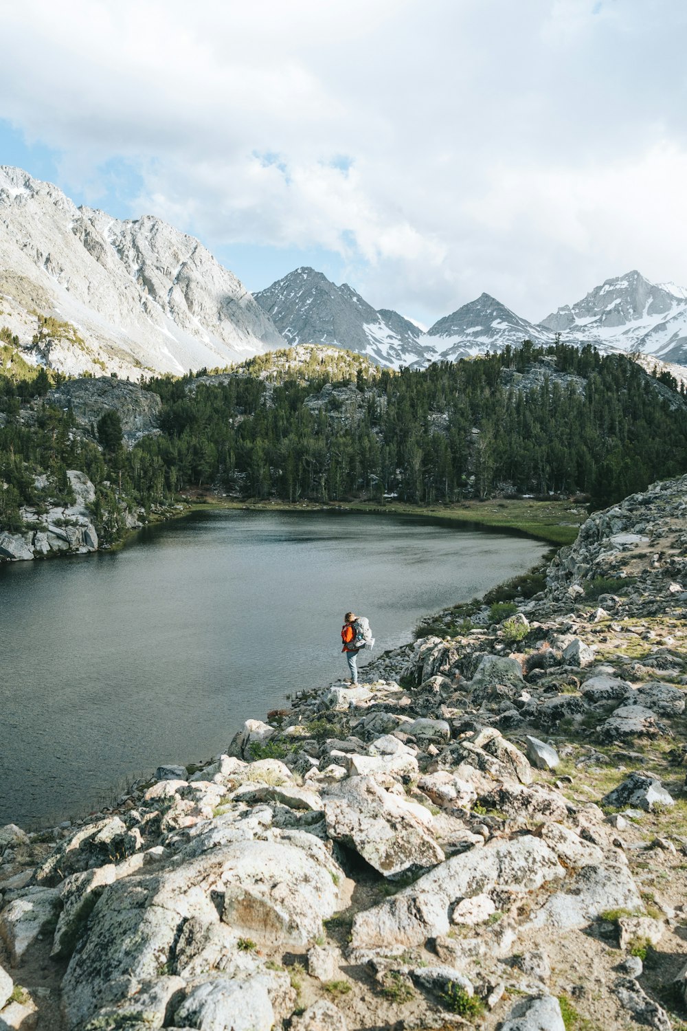 person in red shirt standing on rocky shore near lake and mountains during daytime