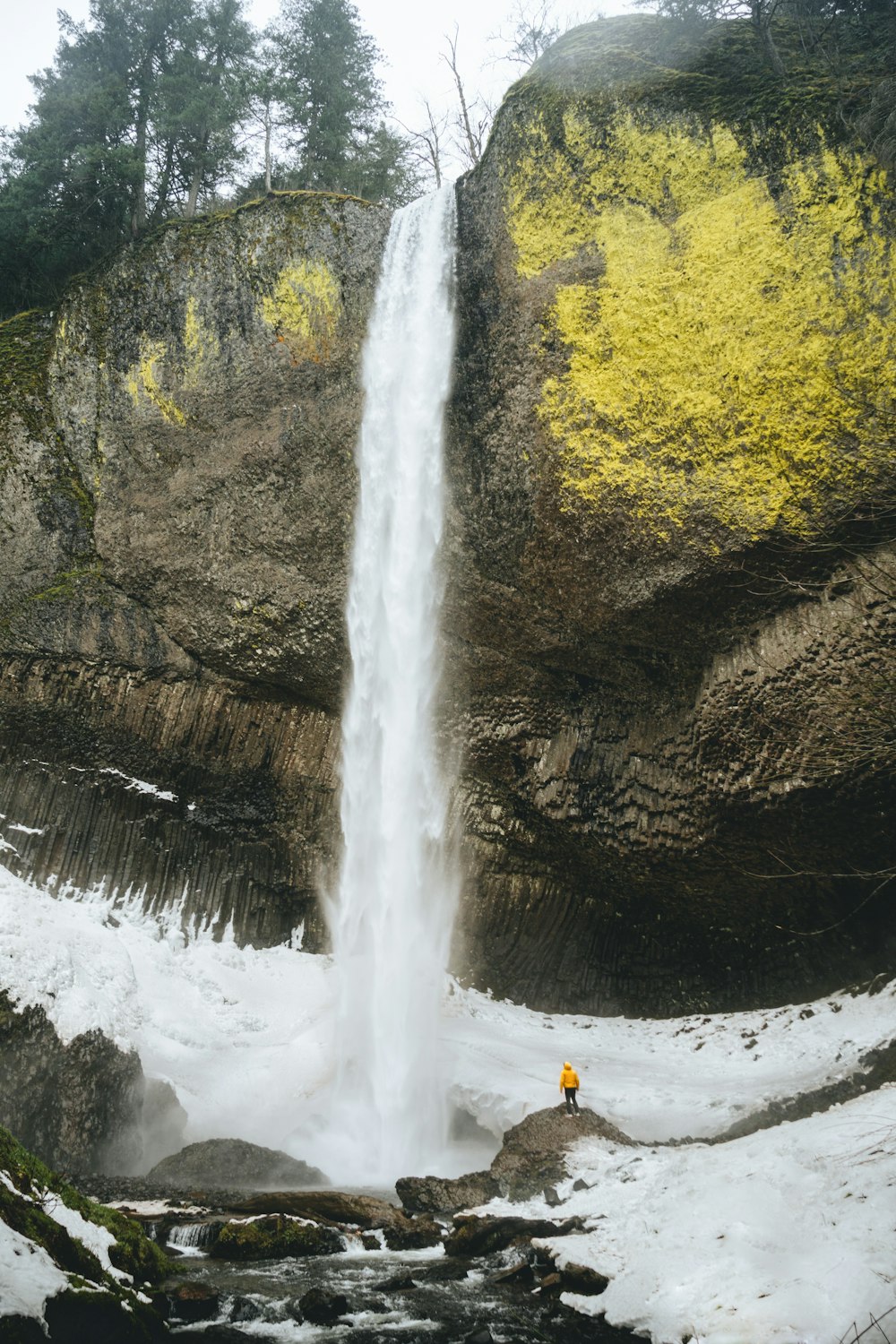 person in orange jacket standing on rock near waterfalls during daytime