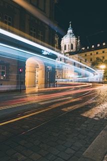 A night scene of a city street with light trails from passing vehicles, showcasing long exposure photography. An ornate, illuminated historical building with a large dome sits in the background, and the cobblestone streets are dimly lit, adding to the urban landscape.