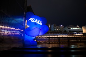 A large blue inflatable bear-like figure with the word 'PEACE' displayed on its chest stands illuminated against a modern metallic building during the night. The cityscape in the background features buildings with lit windows, adding to the urban atmosphere.