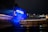 A large blue inflatable bear-like figure with the word 'PEACE' displayed on its chest stands illuminated against a modern metallic building during the night. The cityscape in the background features buildings with lit windows, adding to the urban atmosphere.