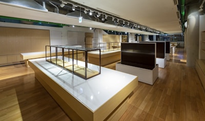 A large, spacious interior with wooden flooring and modern design elements. The room features long white and wooden display tables with metallic frames, possibly used for showcasing items. There are overhead track lights evenly distributed across the ceiling and various shelving units along the walls.
