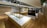 A large, spacious interior with wooden flooring and modern design elements. The room features long white and wooden display tables with metallic frames, possibly used for showcasing items. There are overhead track lights evenly distributed across the ceiling and various shelving units along the walls.