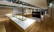 A large, spacious interior with wooden flooring and modern design elements. The room features long white and wooden display tables with metallic frames, possibly used for showcasing items. There are overhead track lights evenly distributed across the ceiling and various shelving units along the walls.