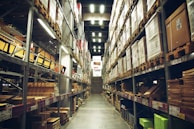 A spacious warehouse aisle flanked by tall shelves filled with various types of boxed goods and merchandise. The aisle has a concrete floor and is illuminated by bright overhead lighting. A large price sign is visible in the distance, indicating an organized retail or storage space.