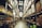 A spacious warehouse aisle flanked by tall shelves filled with various types of boxed goods and merchandise. The aisle has a concrete floor and is illuminated by bright overhead lighting. A large price sign is visible in the distance, indicating an organized retail or storage space.