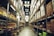 A spacious warehouse aisle flanked by tall shelves filled with various types of boxed goods and merchandise. The aisle has a concrete floor and is illuminated by bright overhead lighting. A large price sign is visible in the distance, indicating an organized retail or storage space.