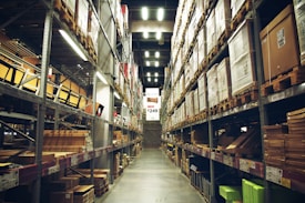 A spacious warehouse aisle flanked by tall shelves filled with various types of boxed goods and merchandise. The aisle has a concrete floor and is illuminated by bright overhead lighting. A large price sign is visible in the distance, indicating an organized retail or storage space.