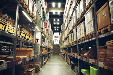 A spacious warehouse aisle flanked by tall shelves filled with various types of boxed goods and merchandise. The aisle has a concrete floor and is illuminated by bright overhead lighting. A large price sign is visible in the distance, indicating an organized retail or storage space.