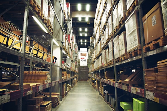 A spacious warehouse aisle flanked by tall shelves filled with various types of boxed goods and merchandise. The aisle has a concrete floor and is illuminated by bright overhead lighting. A large price sign is visible in the distance, indicating an organized retail or storage space.