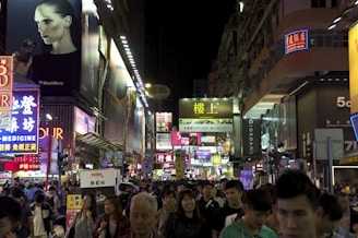 A bustling city street at night is filled with people and vibrant, illuminated advertisements. The street is lined with numerous colorful signs in various languages, showcasing brands and businesses. The atmosphere is energetic, reflecting a busy urban environment.