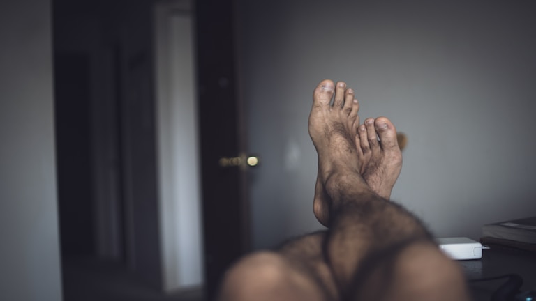A pair of feet with slightly dirty soles and unkempt toenails are visible, suggesting rest or relaxation. The surrounding room is dimly lit with a partially open door in the background, and a small white device sits on a surface to the right.