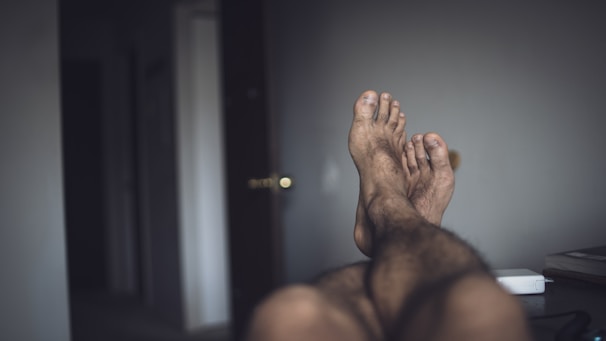 A pair of feet with slightly dirty soles and unkempt toenails are visible, suggesting rest or relaxation. The surrounding room is dimly lit with a partially open door in the background, and a small white device sits on a surface to the right.