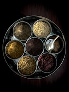 A metal tray holds seven small bowls filled with a variety of spices and ingredients. The spices include cumin seeds, split yellow lentils, fenugreek seeds, coriander seeds, black mustard seeds, red dried chilies, and a bowl containing garlic cloves and ginger. The arrangement is neatly organized, showing a rich texture and variety of ingredients commonly used in cooking.