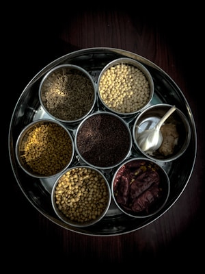 A metal tray holds seven small bowls filled with a variety of spices and ingredients. The spices include cumin seeds, split yellow lentils, fenugreek seeds, coriander seeds, black mustard seeds, red dried chilies, and a bowl containing garlic cloves and ginger. The arrangement is neatly organized, showing a rich texture and variety of ingredients commonly used in cooking.