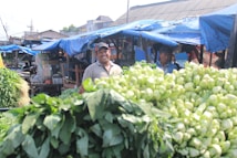 A bustling outdoor market scene with produce displayed in the foreground, including leafy greens and other vegetables. Two men stand behind the stall under blue tarpaulin, one smiling, suggesting a friendly and lively atmosphere.