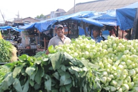 A bustling outdoor market scene with produce displayed in the foreground, including leafy greens and other vegetables. Two men stand behind the stall under blue tarpaulin, one smiling, suggesting a friendly and lively atmosphere.