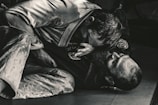 Two individuals are engaged in a Brazilian Jiu-Jitsu match, wearing traditional gis. One person is positioned on top, applying pressure with their upper body, while the other is on their back, appearing to defend or counter the move. The scene is captured in a dimly lit environment, emphasizing the intense physical engagement.