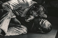 Two individuals are engaged in a Brazilian Jiu-Jitsu match, wearing traditional gis. One person is positioned on top, applying pressure with their upper body, while the other is on their back, appearing to defend or counter the move. The scene is captured in a dimly lit environment, emphasizing the intense physical engagement.
