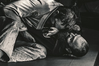Two individuals are engaged in a Brazilian Jiu-Jitsu match, wearing traditional gis. One person is positioned on top, applying pressure with their upper body, while the other is on their back, appearing to defend or counter the move. The scene is captured in a dimly lit environment, emphasizing the intense physical engagement.