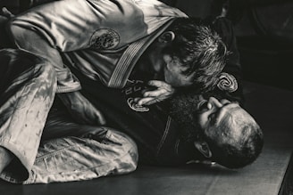 Two individuals are engaged in a Brazilian Jiu-Jitsu match, wearing traditional gis. One person is positioned on top, applying pressure with their upper body, while the other is on their back, appearing to defend or counter the move. The scene is captured in a dimly lit environment, emphasizing the intense physical engagement.