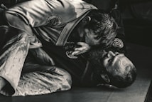 Two individuals are engaged in a Brazilian Jiu-Jitsu match, wearing traditional gis. One person is positioned on top, applying pressure with their upper body, while the other is on their back, appearing to defend or counter the move. The scene is captured in a dimly lit environment, emphasizing the intense physical engagement.