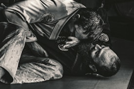 Two individuals are engaged in a Brazilian Jiu-Jitsu match, wearing traditional gis. One person is positioned on top, applying pressure with their upper body, while the other is on their back, appearing to defend or counter the move. The scene is captured in a dimly lit environment, emphasizing the intense physical engagement.