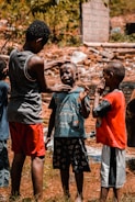A group of young boys are gathered outdoors in a rustic setting. One boy is applying mud or face paint to another boy's face, while a third boy watches and holds his hand to his face.