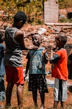 A group of young boys are gathered outdoors in a rustic setting. One boy is applying mud or face paint to another boy's face, while a third boy watches and holds his hand to his face.