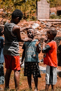 A group of young boys are gathered outdoors in a rustic setting. One boy is applying mud or face paint to another boy's face, while a third boy watches and holds his hand to his face.