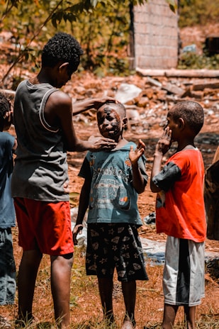 A group of young boys are gathered outdoors in a rustic setting. One boy is applying mud or face paint to another boy's face, while a third boy watches and holds his hand to his face.