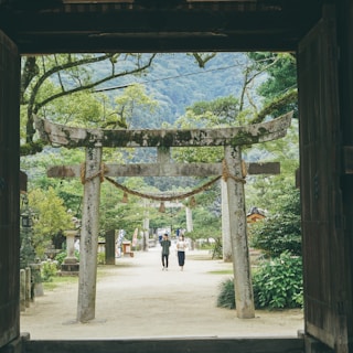 An ancient stone Torii gate stands at the entrance of a lush, green garden path. A couple walks beneath it, surrounded by vibrant trees and foliage. In the background, tall mountains rise under a cloudy sky. Traditional Japanese architecture elements are subtly visible, adding a cultural essence to the serene landscape.
