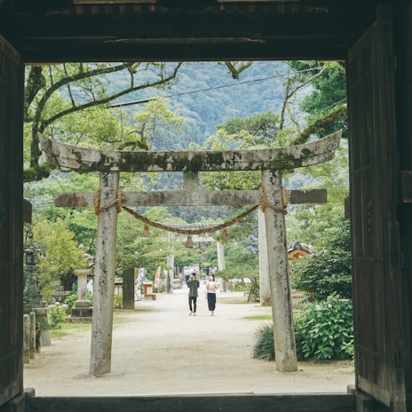 An ancient stone Torii gate stands at the entrance of a lush, green garden path. A couple walks beneath it, surrounded by vibrant trees and foliage. In the background, tall mountains rise under a cloudy sky. Traditional Japanese architecture elements are subtly visible, adding a cultural essence to the serene landscape.