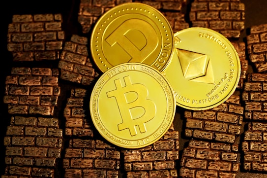 Three golden cryptocurrency coins are displayed on a textured surface resembling small brown bricks. The coins represent Bitcoin, Dogecoin, and Ethereum, each bearing their distinct symbols and inscriptions.