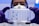 A person wearing a white lab coat, safety goggles, and a protective mask is holding a transparent well plate filled with blue liquid under a bright light. The person is also wearing purple gloves.