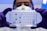 A person wearing a white lab coat, safety goggles, and a protective mask is holding a transparent well plate filled with blue liquid under a bright light. The person is also wearing purple gloves.