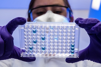 A person wearing a white lab coat, safety goggles, and a protective mask is holding a transparent well plate filled with blue liquid under a bright light. The person is also wearing purple gloves.
