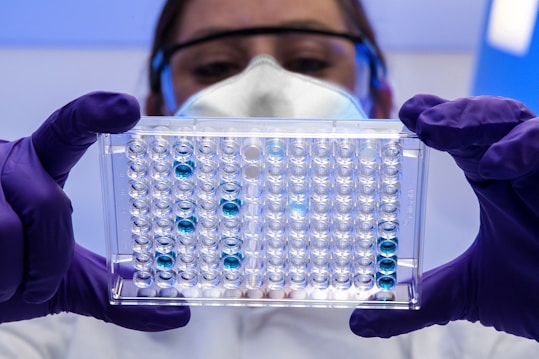 A person wearing a white lab coat, safety goggles, and a protective mask is holding a transparent well plate filled with blue liquid under a bright light. The person is also wearing purple gloves.