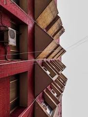 A multi-story building facade with red brickwork and repetitive triangular-shaped concrete structures jutting outwards. Several windows and air conditioning units are visible between the triangles, and there are electrical wires running parallel to the building.