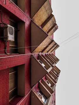 A multi-story building facade with red brickwork and repetitive triangular-shaped concrete structures jutting outwards. Several windows and air conditioning units are visible between the triangles, and there are electrical wires running parallel to the building.