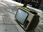 A vintage television set is placed on a paved sidewalk in an outdoor setting. The screen is off, and it shows a bulky design with dials and buttons typical of older television models. In the background, there is a tree with shadows cast on the pavement and a person walking along a street with shops.