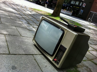 A vintage television set is placed on a paved sidewalk in an outdoor setting. The screen is off, and it shows a bulky design with dials and buttons typical of older television models. In the background, there is a tree with shadows cast on the pavement and a person walking along a street with shops.