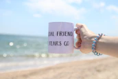 Smiling hands holding a coral-colored mug with a beach scene in the background.