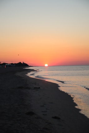 A serene beach scene at sunset with the sun setting on the horizon. The sky is awash with warm hues of orange and pink, reflecting on the calm sea. Silhouetted structures and birds are visible in the distance along the shoreline.