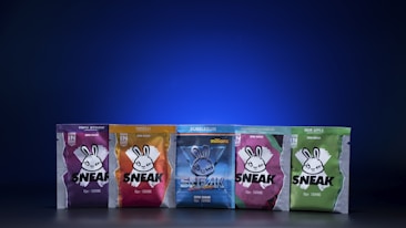 Five brightly colored packages of Sneak energy drink powder are displayed against a dark blue gradient background. Each package features a distinct color and design, with a stylized rabbit logo and bold text. The flavors include Purple Storm, Tropikilla, Bubblegum Millions, Strawberry Watermelon, and Sour Apple.