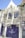 A quaint white house with vibrant purple windows and door, adorned with lush wisteria vines. Sunlight casts shadows of the wisteria on the facade. There is a sign indicating 'Number 11 Exclusive Guesthouse'. Greenery climbs the walls, and several road signs are attached to the building.