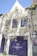 A quaint white house with vibrant purple windows and door, adorned with lush wisteria vines. Sunlight casts shadows of the wisteria on the facade. There is a sign indicating 'Number 11 Exclusive Guesthouse'. Greenery climbs the walls, and several road signs are attached to the building.