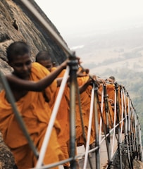 A group of people wearing saffron robes ascends a steep and rocky path protected by a handrail. The setting is elevated with a view of the landscape below, suggesting they are climbing a significant height. The vibrant orange color of the robes stands out against the natural surroundings.