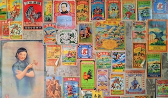 A collage of vintage Chinese posters and advertisements with colorful and intricate designs. The posters feature a variety of images including traditional symbols, animals like roosters and dragons, and a portrait of a woman in a traditional dress holding flowers. The images are arranged tightly together, covering a wall, with an aged and nostalgic aesthetic.