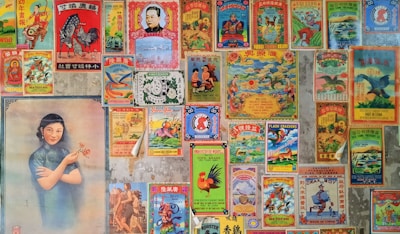 A collage of vintage Chinese posters and advertisements with colorful and intricate designs. The posters feature a variety of images including traditional symbols, animals like roosters and dragons, and a portrait of a woman in a traditional dress holding flowers. The images are arranged tightly together, covering a wall, with an aged and nostalgic aesthetic.